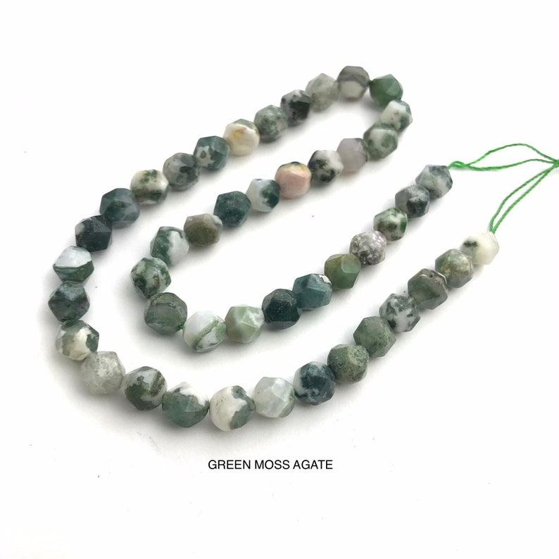 May include: Strand of faceted green moss agate beads. The beads have a mix of green, white, and gray colors. The text "GREEN MOSS AGATE" is printed at the bottom of the image. The beads are strung on a green thread.