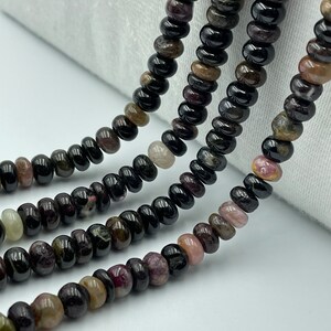 May include: A strand of black tourmaline beads with hints of pink, green, and brown. The beads are round and smooth, and they are strung on a clear thread.