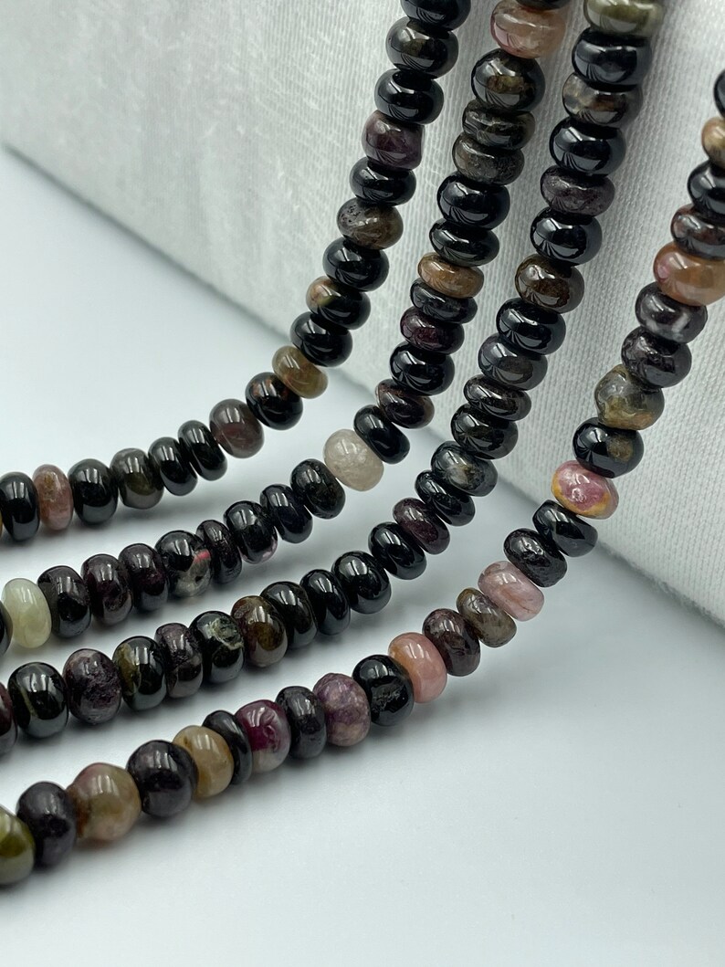 May include: A strand of black tourmaline beads with hints of pink, green, and brown. The beads are round and smooth, and they are strung on a clear thread.