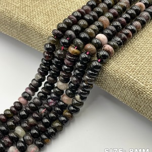 May include: A strand of black tourmaline beads with green and pink inclusions. The beads are 8mm in size and 4-5mm thick.  TOURMALINE is written at the top of the image. SIZE: 8MM and THICKNESS: 4-5MM are written at the bottom of the image.