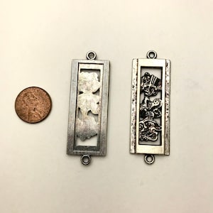 May include: Two silver-toned metal charms with intricate designs. The charm on the left features a floral design, while the charm on the right features a design of three figures. Both charms have a rectangular shape with a loop at the top.