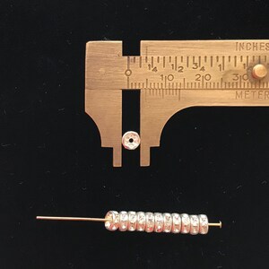 Puede incluir: Primer plano de una cuenta de metal plateado con un di&aacute;metro de 1,27 cm, medida con un calibre. La cuenta est&aacute; rodeada por una fila de cuentas plateadas m&aacute;s peque&ntilde;as en un alambre fino de oro.