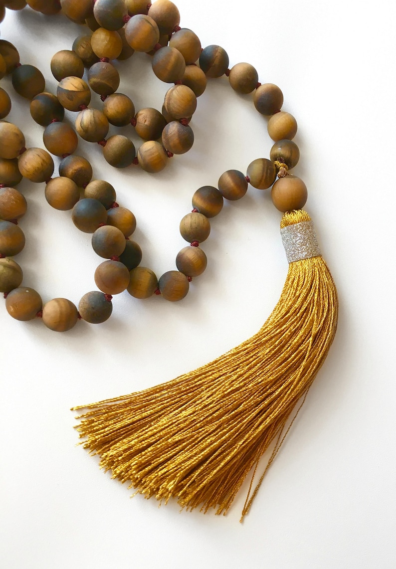 May include: A brown tiger's eye gemstone mala with a gold tassel. The mala is made of round beads and has a red string connecting the beads.