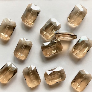 May include: A collection of twelve rectangular glass beads in a light brown color. The beads have a faceted design and are arranged on a white surface.