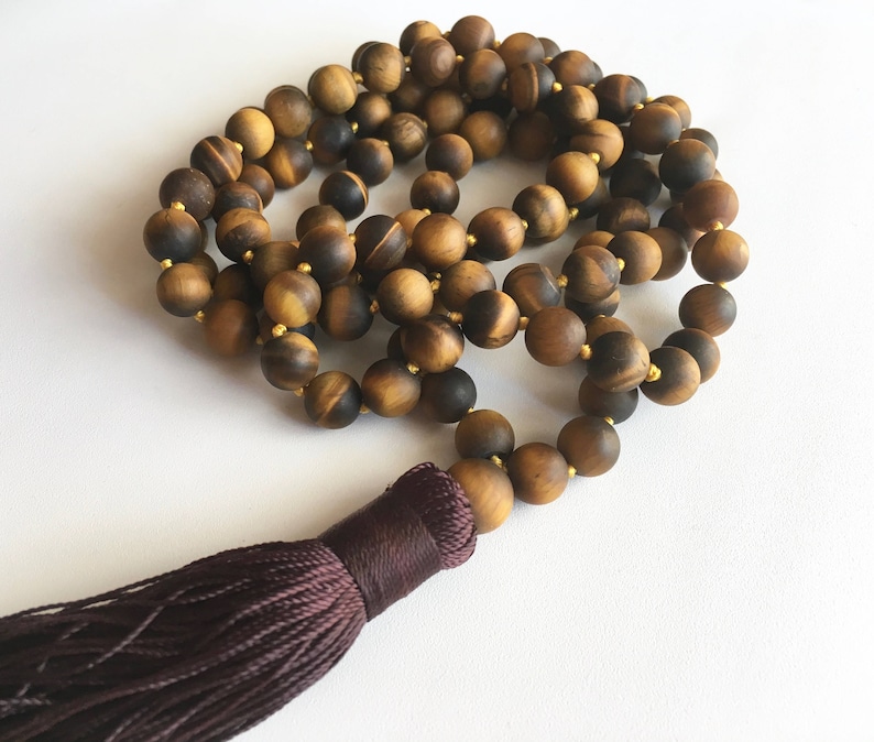 May include: A brown and gold tiger's eye mala bead necklace with a dark purple tassel. The necklace is made of round beads and is approximately 108 beads long.