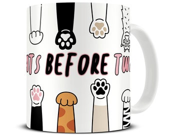 May include: White ceramic mug featuring a design of various cat paws in black, tan, and pink. The mug has the text "CATS BEFORE TW" in black and pink lettering. The mug has a white handle.