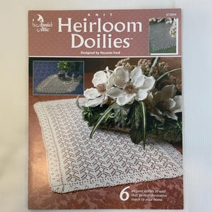May include: A book cover for "Heirloom Doilies" by Annie's Attic. The cover features a white lace doily and a floral arrangement. The text reads "6 elegant doilies to add that perfect decorative touch to your home."