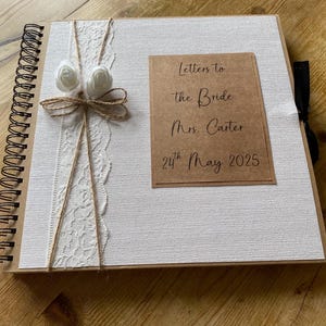 Puede incluir: Un álbum de boda con una cubierta blanca texturizada y un borde marrón. La cubierta está decorada con encaje blanco, hilo y dos rosas blancas. Una tarjeta marrón con el texto "Letters to the Bride Mrs. Carter 24th May 2025" está adherida.