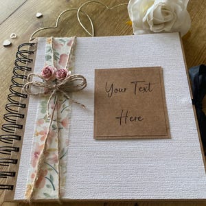 May include: A white scrapbook with a spiral binding, decorated with a floral ribbon and twine bow. The cover has a brown label with the text "Your Text Here". A white rose and gold heart-shaped wire are also present.