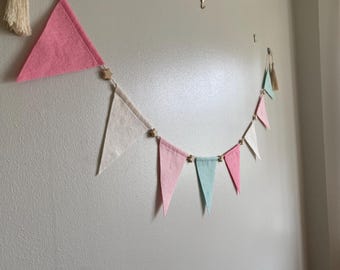 Könnte beinhalten: Eine dekorative Wimpelkette mit abwechselnden dreieckigen Stoffwimpeln in den Farben Rosa, Creme und hellem Türkis. Die Wimpel sind an einer Schnur befestigt, die mit kleinen, hellen, sternförmigen Holzperlen und Quasten verziert ist.