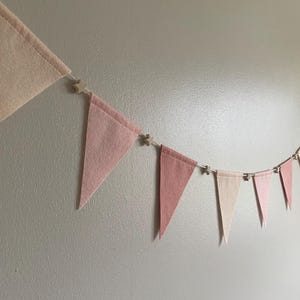 May include: A garland of triangular flags in various shades of pink and cream, connected by small wooden star-shaped embellishments. The banner is displayed against a neutral wall.