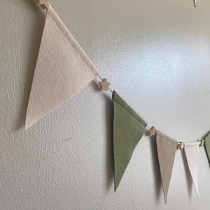 May include: A felt banner featuring triangular flags in beige and green, connected by wooden star-shaped beads. The banner is displayed against a light grey wall, offering a simple, decorative touch.