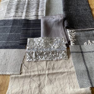Op de afbeelding: Een verzameling stofstalen in verschillende tinten grijs, beige en wit. De stalen bevatten patronen, texturen en pailletten. Sommige stoffen hebben strepen, geometrische ontwerpen of bloempatronen. De stoffen zijn gerangschikt op een houten oppervlak.