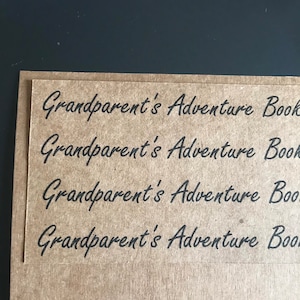Könnte beinhalten: Nahaufnahme einer Kraftpapierkarte mit dem schwarzen Schriftzug "Grandparent's Adventure Book". Die Karte ist auf einer weiteren Kraftpapierkarte angeordnet. Der Hintergrund ist eine dunkelgraue Oberfläche.