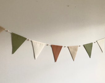 Può includere: Una ghirlanda di bandierine triangolari in tessuto di colore beige, verde e marrone. Le bandierine sono infilate su uno spago bianco e sono appese a un muro bianco.