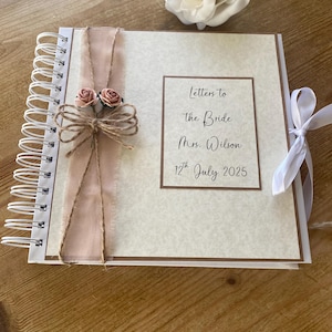 Może przedstawiać: Biała i brązowa książka w oprawie spiralnej z beżową okładką. Na okładce widnieje tekst "Letters to the Bride Mrs. Wilson 17th July 2025". Książka jest ozdobiona różową wstążką, dwoma małymi różowymi kwiatami i sznurkiem.