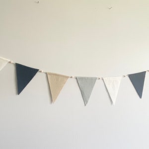 Könnte beinhalten: Eine Girlande aus dreieckigen Stofffahnen in den Farben Weiß, Beige, Grau und Schwarz mit Quasten an jedem Ende. Die Fahnen sind an einer weißen Schnur mit kleinen Holzperlen dazwischen befestigt.