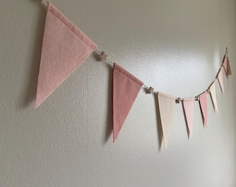 May include: A pink felt banner with triangle flags in varying shades of pink and cream. The flags are strung together with a white string and small star-shaped wooden clips. The banner is hanging against a light gray wall.