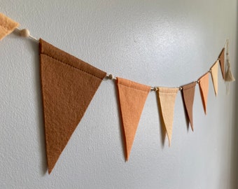 Op de afbeelding: Een slinger van vlaggetjes van vilt in tinten bruin, oranje en crème, geregen aan wit touw met houten kralen.
