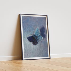 Puede incluir: Obra de arte enmarcada con una mariposa azul sobre un fondo azul claro texturizado. La mariposa tiene alas detalladas con tonos de azul y un cuerpo oscuro. La obra está en un marco negro, apoyada contra una pared blanca y un suelo de madera.