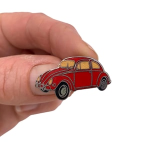 May include: A red enamel pin in the shape of a classic car. The pin features a detailed design with black tires, silver accents, and yellow-tinted windows. The car is held in a hand against a white background.