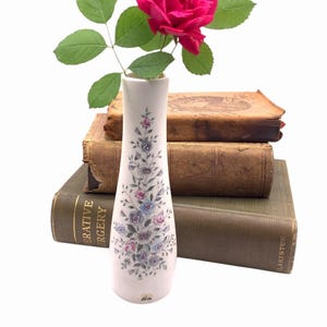 May include: A white ceramic vase with a floral pattern holds a vibrant pink rose with green leaves. The vase sits atop a stack of old books with worn, brown covers, creating a vintage aesthetic.