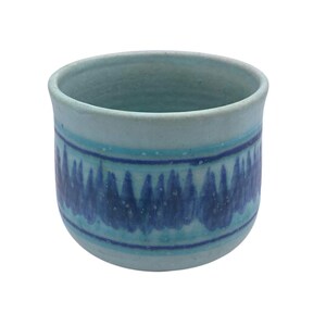 May include: A ceramic cup with a light blue base color, featuring a horizontal band of dark blue, jagged, flame-like patterns encircling the middle. The rim is smooth and rounded, and the overall finish of the vessel appears matte and textured.