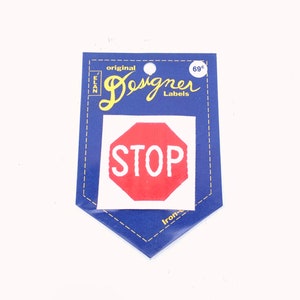 Op de afbeelding: Strijk-op patch met een rood stopteken met witte letters. De patch is op een blauwe achtergrond met de tekst "original Designer Labels" en "69¢".