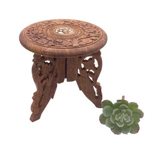 May include: A small, intricately carved wooden plant stand with a round top and three decorative legs. The stand is brown and features floral patterns. A green succulent plant is next to the stand.