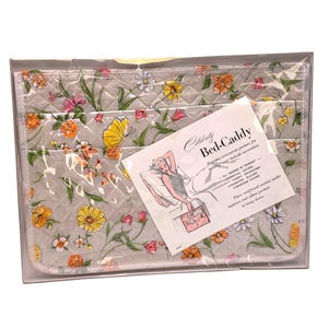 May include: A quilted gray bed caddy featuring a colorful floral pattern with yellow, orange, and pink flowers and green leaves. The product is packaged in a clear plastic sleeve with an attached white card labeled Celebrity Bed-Caddy.