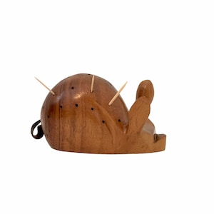 Puede incluir: Un porta palillos de dientes de madera con forma de cactus, con un acabado marrón. El porta palillos tiene varios agujeros para palillos y está decorado con pequeños puntos negros.