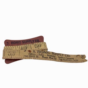 Peut inclure: Un ruban à mesurer en tissu beige vintage enroulé autour d'une bobine en carton rouge foncé. Le ruban présente des chiffres noirs imprimés et le texte S. HONIG SUPPLY CO. Clothiers Tailors Dressmakers Supplies & Equipment 1844 GEARY ST. Phone: West 7147 SAN FRANCISCO CALIF.