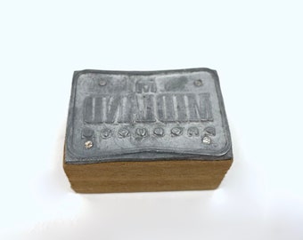 Puede incluir: Un sello de metal plateado con la palabra "MOUNTAIN" en relieve. El sello está montado sobre una base de madera.