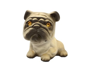 Può includere: Una piccola statuetta di ceramica beige di un carlino con occhi marroni. La statuetta è seduta con la testa leggermente inclinata di lato.