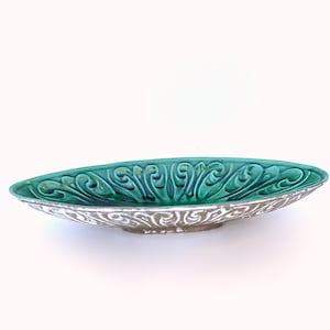 May include: A ceramic serving dish with an oval shape and a green glaze. The dish has a raised design of stylized leaves and swirls.