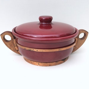 May include: A red ceramic casserole dish with wooden handles and copper accents. The dish has a lid and is perfect for serving a meal.