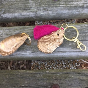 May include: Two gold oyster shell keychains. One keychain has a pink tassel and a gold metal ring. The other keychain has a gold metal ring.