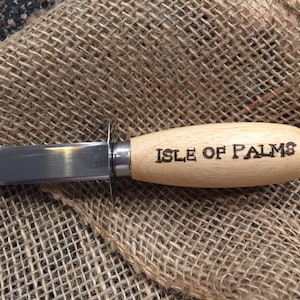 May include: A stainless steel oyster knife with a light-colored wooden handle. The handle features the text ISLE OF PALMS burned into the wood. The knife has a short, pointed blade and a small metal guard between the blade and the handle.