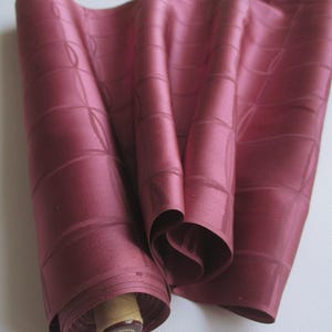 May include: A roll of deep red fabric with a grid-like design. The fabric is rolled up, revealing a wooden spool. The material has a slight sheen, possibly silk or satin. The fabric is draped, showing its texture and colour. The overall effect is elegant.