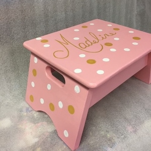 May include: A pink wooden step stool with white and gold polka dots. The stool is personalized with the name "Madeline" in gold lettering.
