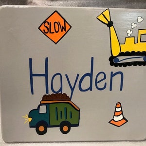 May include: A gray wooden box with a blue inscription that says "Hayden". The box is decorated with a yellow construction vehicle, a green dump truck, a yellow and orange traffic cone, and a yellow sign that says "SLOW".