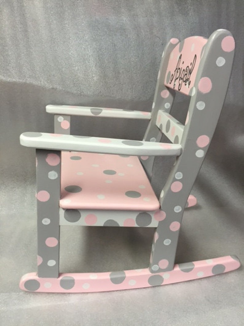 May include: A gray and pink rocking chair with white polka dots. The chair is personalized with the name "Abigail" on the back.