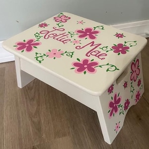 May include: A white wooden step stool with a rectangular top and two steps. The top is decorated with pink flowers and green vines, and the name "Lottie Mae" is written in cursive. The sides also feature floral designs.