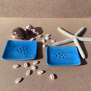 May include: Two blue ceramic soap dishes with drainage holes. The dishes are on a light brown surface with white seashells scattered around them. There is a starfish and two seashells in the background.