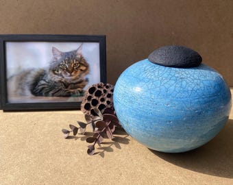 Könnte beinhalten: Eine blaue Keramikurne mit schwarzem Deckel steht neben einem gerahmten Foto einer Katze. Die Urne hat eine Craquelé-Glasur. Getrocknete Samenkapseln und künstliches Laub sind ebenfalls im Bild. Der Fotorahmen ist schwarz.