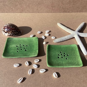 May include: Two green ceramic soap dishes with drainage holes. The dishes are on a brown surface with white seashells scattered around them.
