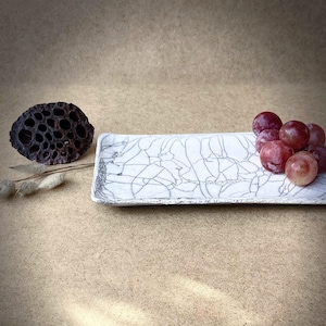 Può includere: Un vassoio da portata in ceramica bianca con una glassa craquelé e un design a linee bianche e nere. Un grappolo d'uva rossa è sul vassoio.