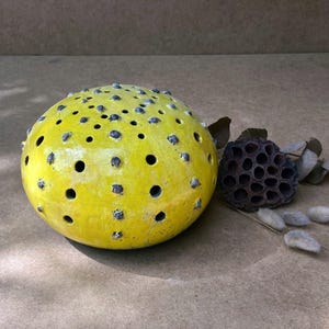 May include: A yellow ceramic sphere with black dots and holes throughout the surface. The sphere is sitting on a brown surface.