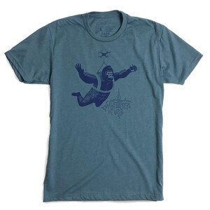 May include: A teal blue t-shirt with a navy blue graphic of a gorilla wearing a parachute and flying over a city skyline with a drone above.