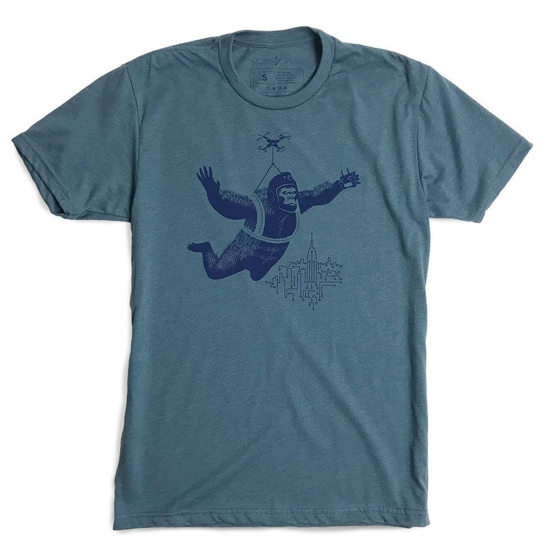 May include: A teal blue t-shirt with a navy blue graphic of a gorilla wearing a parachute and flying over a city skyline with a drone above.